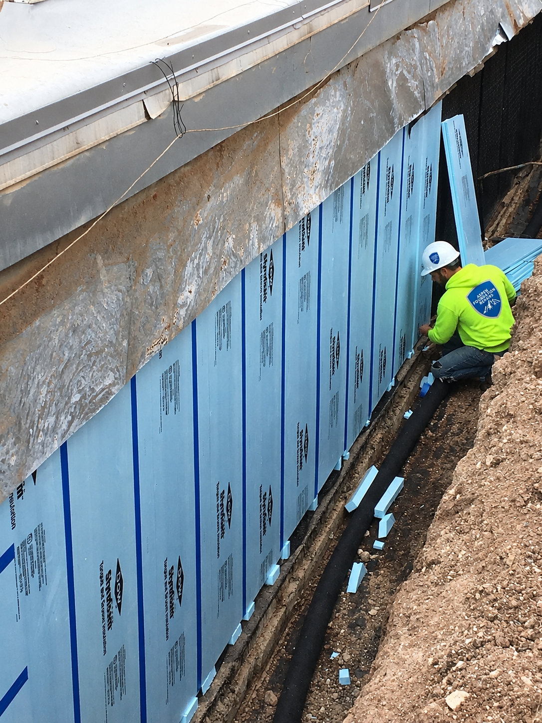 Crew fitting rigid foam board over waterproofing on an excavated foundation wall